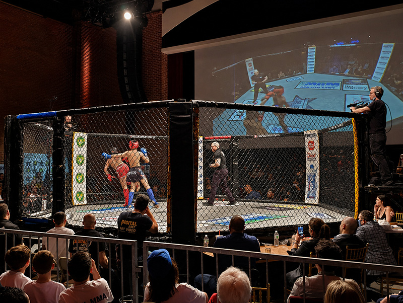 Two fighters competing inside a cage while the live event is displayed on a projection screen
