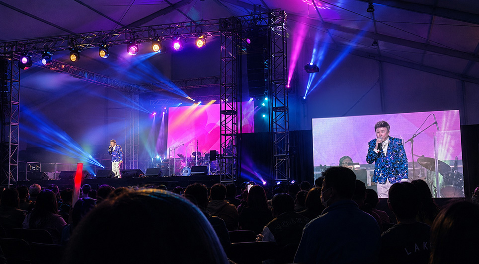 Male singer performing on a brightly lit concert stage with a large video screen